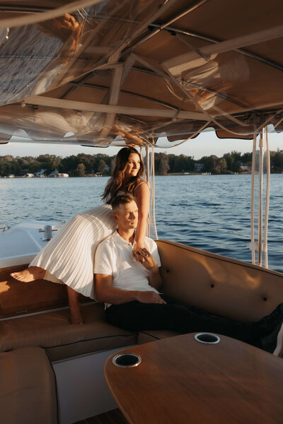 Woman in white dress sits behind man in white polo with her hand on his chest as they look off into the sunset