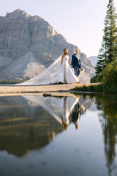 Bride and groom walking hand in hand at Bow Lake in Banff National Park with their reflection visible in the water and Bow Mountain in the background on a warm summer afternoon