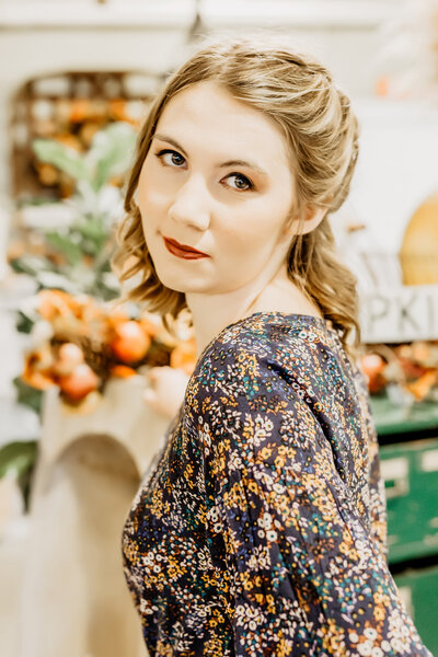 A high school senior girl wearing a floral shirt, standing in a whimsical room, with an orange & green accents, while posing for a south shore ma senior photographer