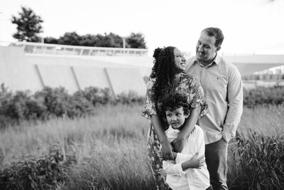 a family with mom and dad looking at each other lovingly while hugging son near the hunters point south park in long island city new york in black and white outdoors
