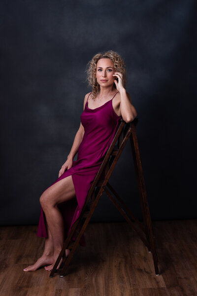 moody portrait of a blonde woman in a silk dress leans casually on a wooden ladder