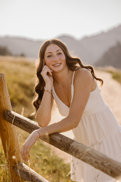 High school senior girl in a white dress leans on a rustic wooden fence at Chautauqua Park in Boulder, Colorado, smiling with golden sunlight and mountains in the background.