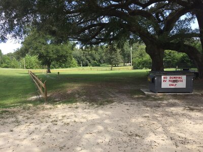A large tree provides shade over a grassy area with a dirt path. To the right, a dumpster bears a sign: "NO DUMPING, RV PARK USE ONLY." A wooden fence outlines part of the open field in the background, reminiscent of Mayo Florida camping near Suwannee River Rendezvous.