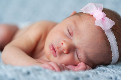 Monroe newborn photographer poses baby peacefully sleeping on a soft blue blanket, wearing a delicate pink bow headband. 