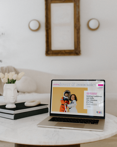 A laptop on a table displays a Sweet Pea Ink Creative web design featuring two children hugging. Nearby are a vase with white tulips, stacked books, and a mirror on the wall in a minimalist room.