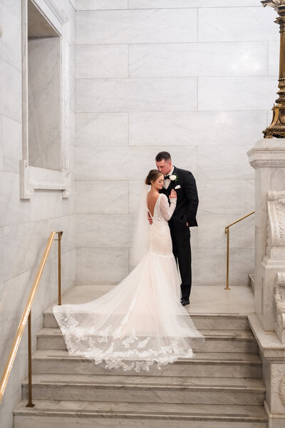 A bride in a flowing white dress and her husband in a black suit hold each other and smile at the camera