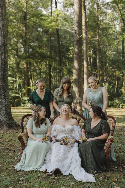 A person in a wedding dress sitting in a chair in a field with their wedding party sitting and standing next to/behind them 