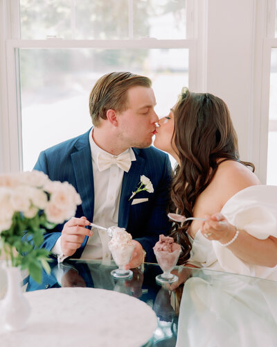A newlywed couple kissing as they hold up spoons of ice cream 
