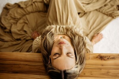 A woman with long blonde hair and a flowing tan dress lies on a wooden surface with her eyes closed.
