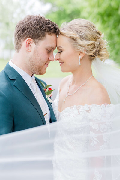 A man and woman stand forehead to forehead as her wedding veil swirls around them.