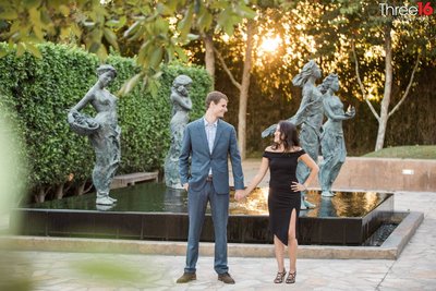 Engaged couple hold hands and look at each other as they pose for engagement photos in front of some statues at Sculpture Garden in Cerritos