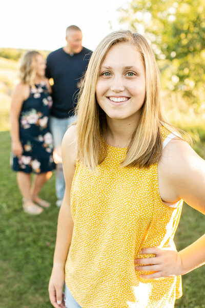 A white teenager wearing a yellow tank top with her family in the background