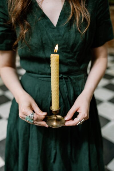 Kassie, the copywriter behind Write by Firelight, holding a lit candle in a moody greenhouse with black and white-tiled checkerboard-style floor at Sekrit Theatre in Austin Texas