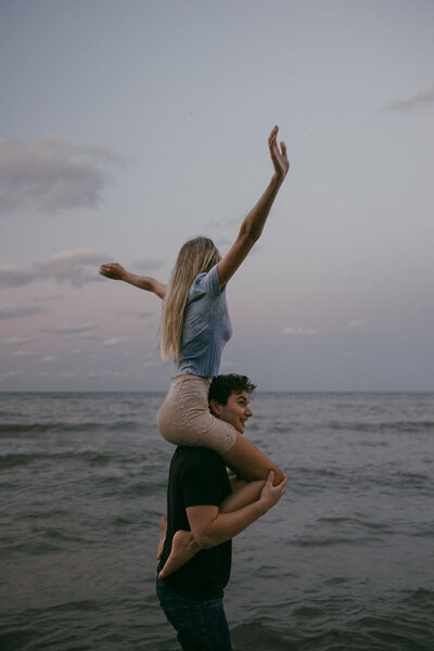 Beautiful dark and moody engagement photography of bride and groom on the beach at sunset in front of the water in Milwaukee, WI at Lake Michigan, the groom is holding the bride on his shoulders and the bride is sticking her hands up in the air. Engagement photos captured by Morgan Ashley Lynn Photography