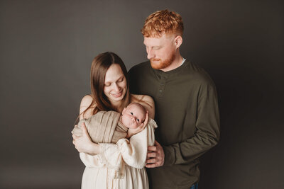 A cozy family moment captured indoors, with a mother and father cradling their newborn baby near a softly lit window. The father gently kisses the mother’s head while she looks down lovingly at their sleeping baby. The natural light enhances the warmth and tenderness of the scene.