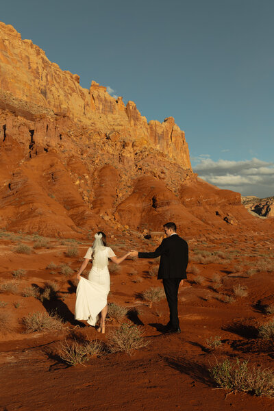 A bride and groom are facing each other, both looking down at the vows which in written in small books. They are in southern Utah, standing in a field of sagebrush and orange dirt with beautiful buttes behind them.