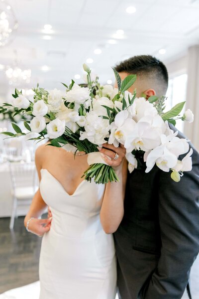 romantic wedding moment where a bride and groom are hidden behind the bride's elegant bouquet. The bouquet features white flowers, including roses, lisianthus, and orchids, with lush green foliage. The bride is wearing a strapless white gown, while the groom is in a dark suit. The background shows a beautifully decorated reception venue with white chairs, floral arrangements, and soft, natural light from large windows, creating a dreamy and elegant atmosphere.