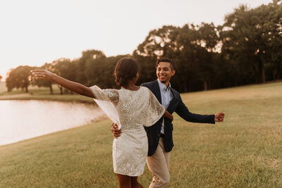 Couple in dress and suit hold each other as they spin  in a field with pond behind them