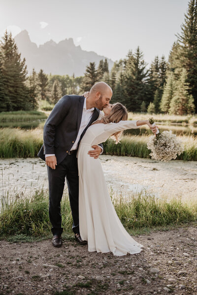 celebratory kiss between bride and groom in front of a pond in the woods with the grand tetons in the distance captured by jackson hole wedding  photographers