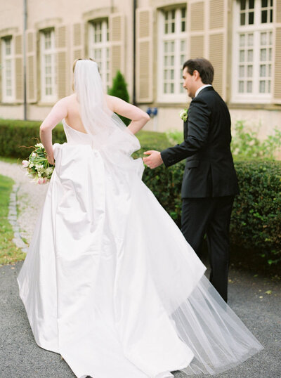 Glen Manor House | A bride in a flowing white gown and veil holds a bouquet, walking beside a groom in a black suit. They are on a path near a building with a hedge.