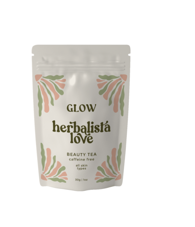 A pouch of Herbalista Love Glow herbal tea displayed against a peach-colored background. The packaging features delicate green and pink leaf patterns around the brand name, emphasizing the product’s natural and organic qualities. The design reflects a fresh, holistic approach to wellness.