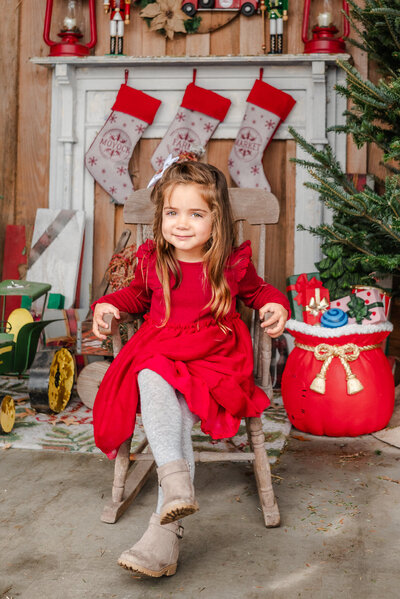 A young girl, wearing a red dress, sits on a child's rocking chair. She is in front of a Christmas tree and a faux fireplace with stockings hung up.