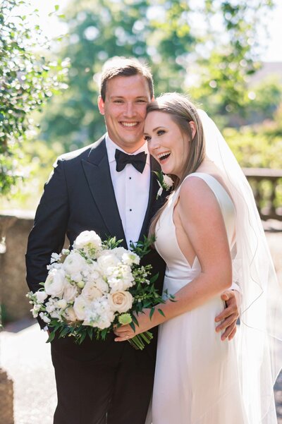 Groom smiles at the camera while holding the waist of his blonde bride