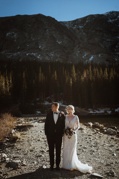 groom and bride are standing next to each other looking out into the distance at an alpine lake in breckenridge. the bride is holding her flowers and she has her hand on the groom's shoulder. there are mountains with snow in the background.