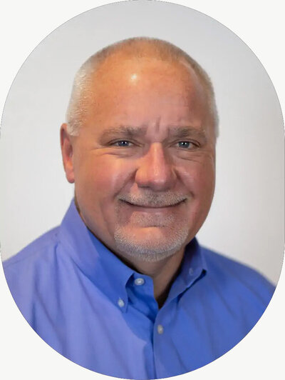 Portrait of a man in a blue button-up shirt, smiling warmly against a light background.