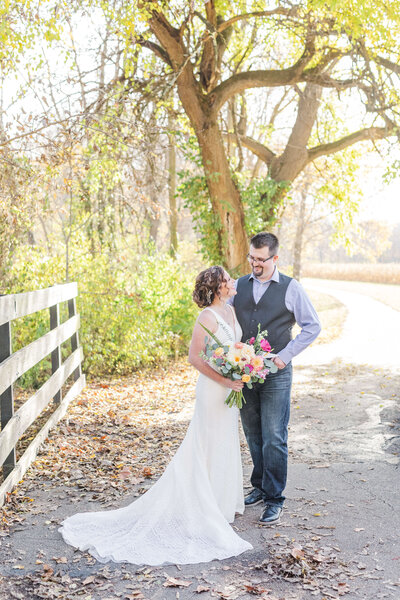 A bride and groom are standing on a walkway in the woods as the golden sun shines through the trees. She is holding her bouquet full of bright pink and orange flowers. They are looking at each other as they have their arms around each other.