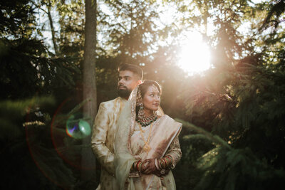 A bride and groom stand amidst lush greenery, illuminated by golden sunlight filtering through the trees, exuding elegance and serenity.