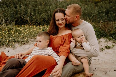 Family of four sitting on the sand at the beach. Mom smiling at Dad.