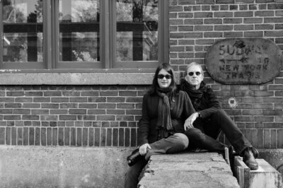 Black and white branding photo of Open Book Music duo Rick and Michele Gedney leaning against brick wall in downtown Cold Spring, NY.