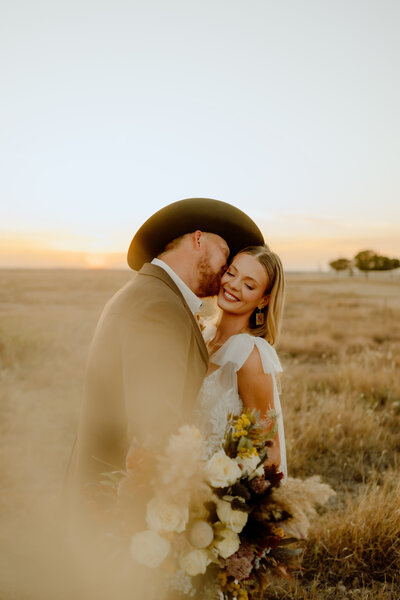 groom kisses his bride at sunset on their wedding day while she holds her bouquet