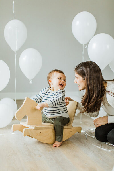 mom helps push son on rocking horse surrounded by balloons for first birthday photo session