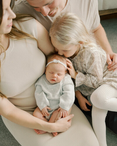 sister giving newborn baby sister soft kiss on the head with mom and dad watching from above