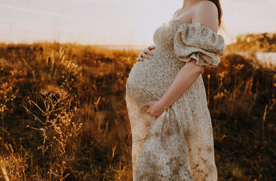 A mom holding her pregnant belling standing in a field of tall brown grass near Grapevine Lake at Murrell Park in Flower Mound, TX