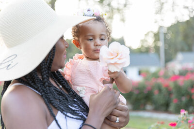 A mother holding a young toddler girl in a sun light rose garden as the toddler admires a soft pink rose she holds in her hand. 