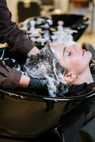 Professional stylist performing scalp detox treatment with foaming cleanser at salon wash station.