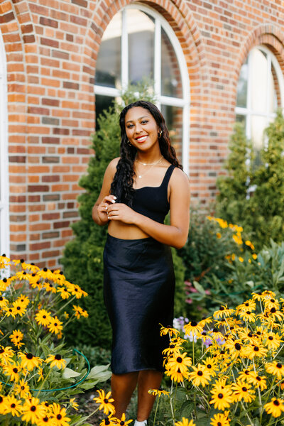 Senior in a black outfit posing in front of a brick wall and yellow flowers
