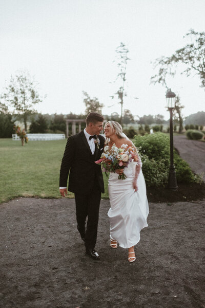 A bride and groom standing outside  RiverCrest wedding venue in Phoenixville, Pa. The groom is smiling down at his bride, while the bride is smiling at the camera. The groom is wearing a maroon and black tux. The bride in a beautiful floral detailed white gown. Phoenixville wedding photographer.