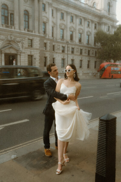 bride standing in front of groom, her back to his chest,  arms wrapped around each other, bride holding the ends of her dress, while the city traffic rushes by with a blurred background together