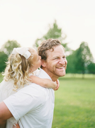 Dad laughs at something his blonde, toddler daughter whispers in his ear as she rides piggyback