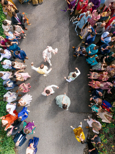 Aerial view of a lively street dance scene with people dressed in colorful traditional outfits. A group is dancing energetically in the center, surrounded by onlookers clapping and cheering along the road.