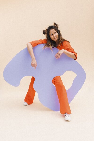 Woman smirks wearing an orange jumpsuit with one leg stretched through a the hold of a purple abstract shape made of wood.