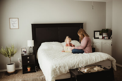 Mom and toddler daughter open a jewelry box together on the bed.