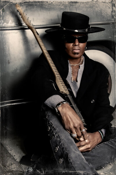 Musician Portrait Jeau James itting against silver guitar guitar leaning on crossed legs wearing black hat and sunglasses Package B