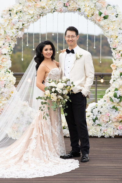 Bride and  groom wedding photo outside standing on a deck at the Trump National Golf Club  overlooking the golf coarse with a giant white rose florial wreath behind them.