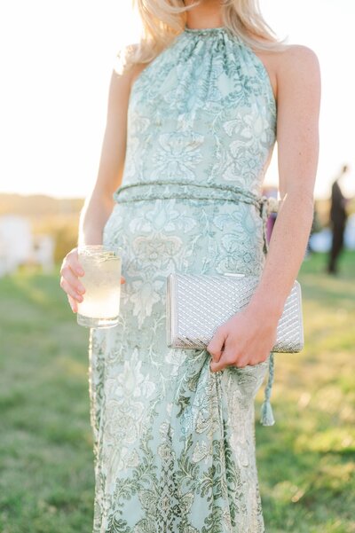 A wedding guest holds a drink and a clutch as the sun sets behind her.