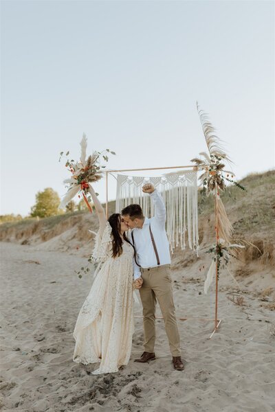 bride and groom celebrating and kissing on a beach in front of their ceremony arch
